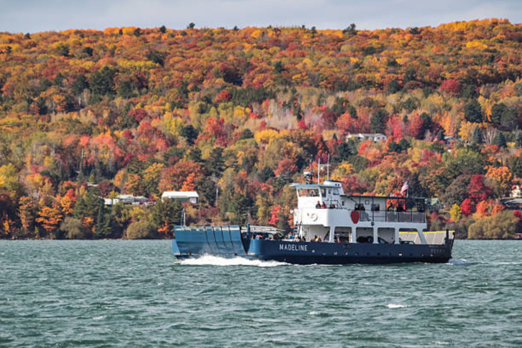 Ferry crossing a Great Lakes channel below a hillside of orange and red color, a scenic stop on Midwest fall foliage trips