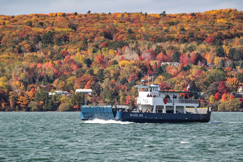Ferry crossing a Great Lakes channel below a hillside of orange and red color, a scenic stop on Midwest fall foliage trips