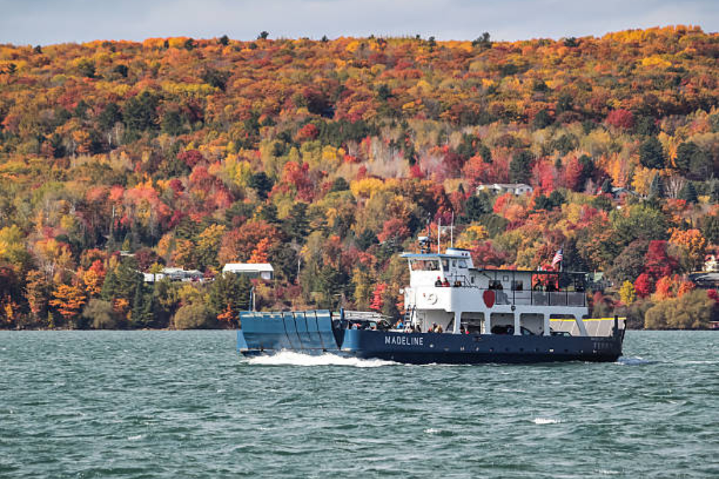 Ferry crossing a Great Lakes channel below a hillside of orange and red color, a scenic stop on Midwest fall foliage trips