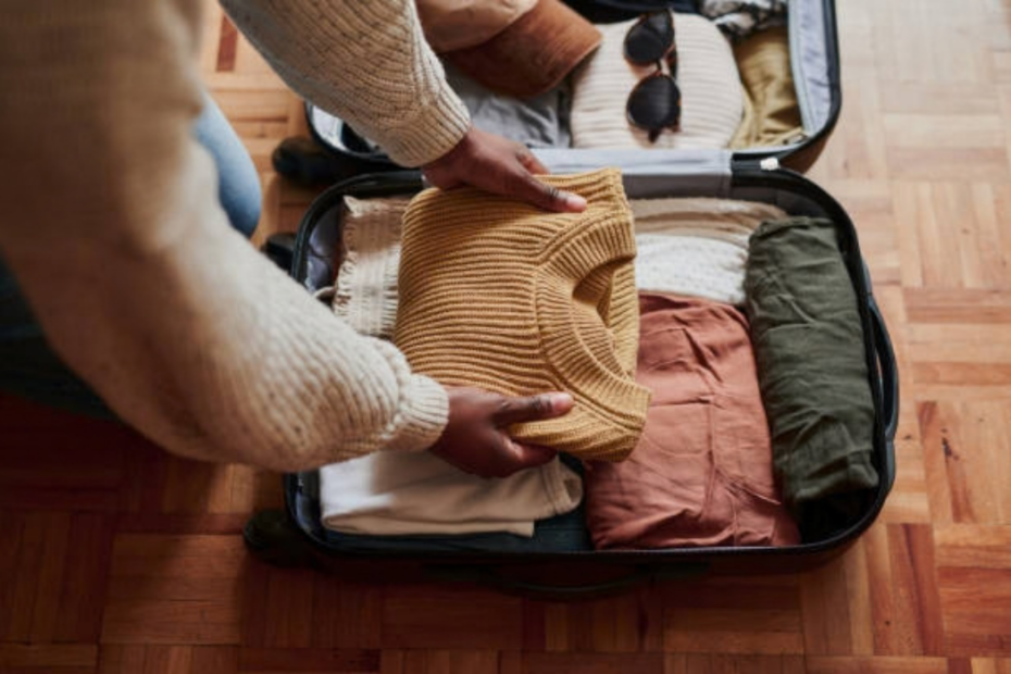 Person packing a mustard knit sweater and other folded clothes into an open suitcase on a wood floor
