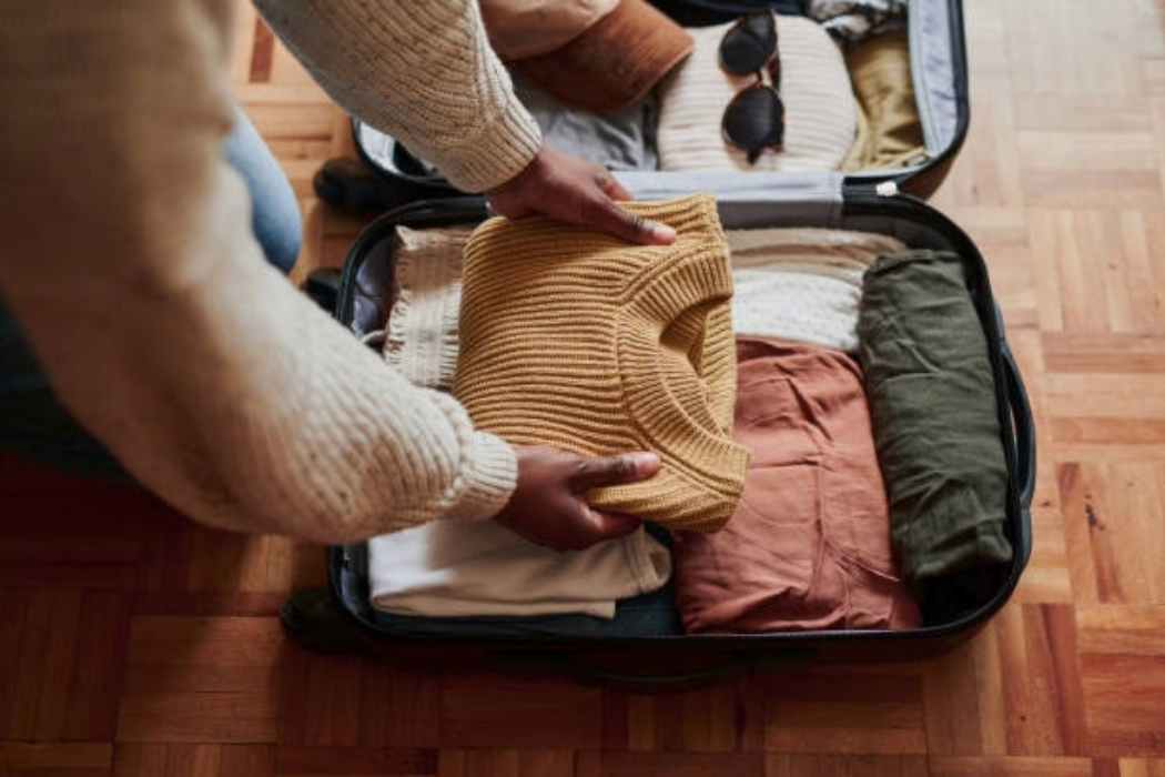 Person packing a mustard knit sweater and other folded clothes into an open suitcase on a wood floor